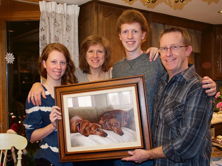 The Fitzpatrick family smiling while holding the framed oil portrait of their two Dachshunds, Maggie and Max.