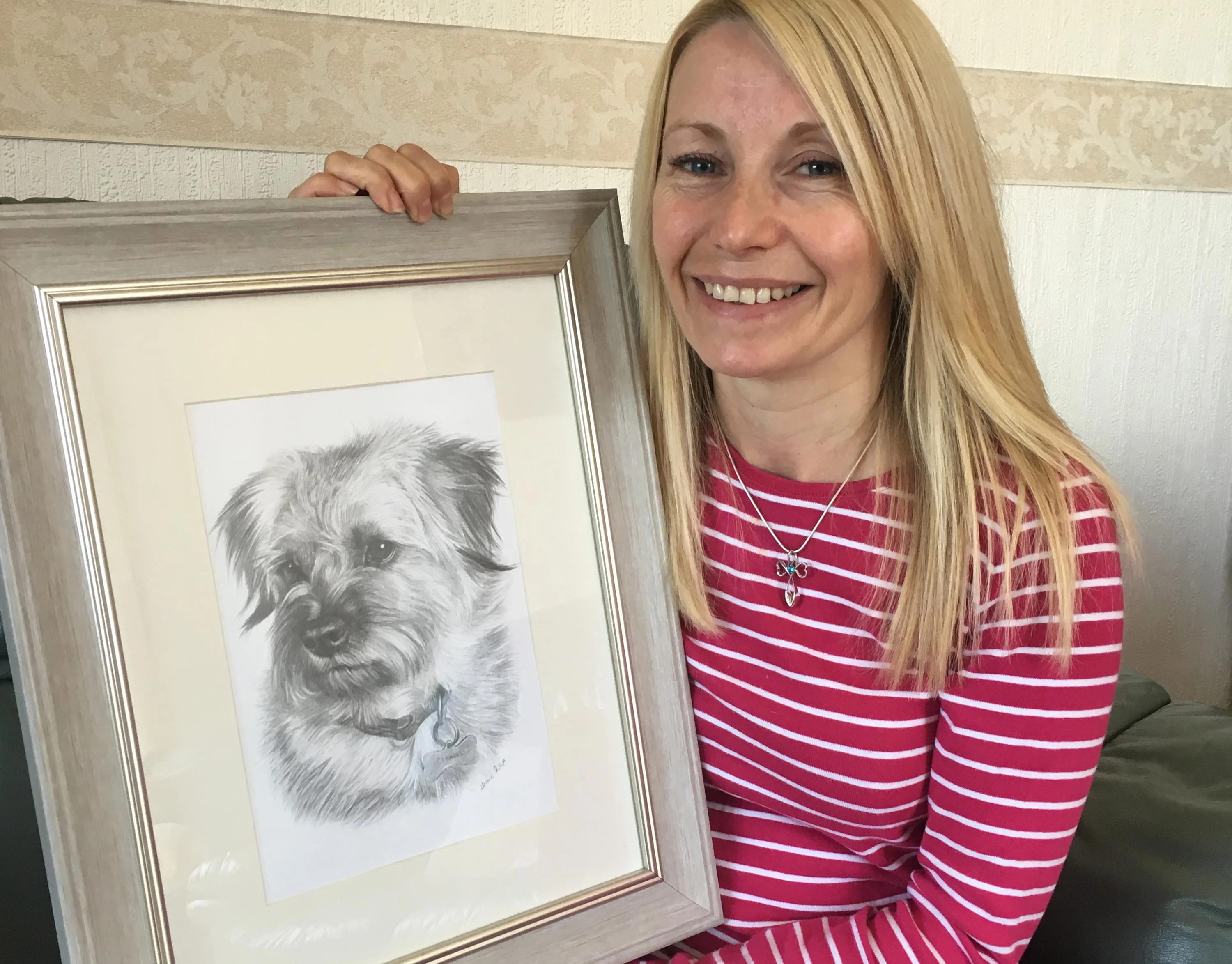 Framed Pencil Portrait of a Border Terrier Cross – Custom Artwork by Melanie Phillips Happy client, Paula, holding a framed pencil portrait of her Border Terrier cross dog, Teasel.