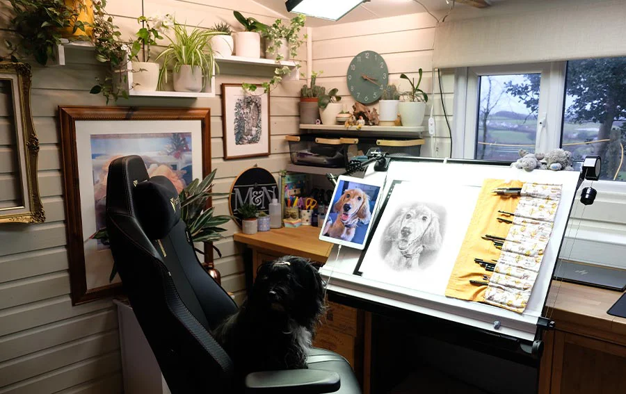 Melanie's organized drawing desk inside the art studio, featuring a tilted drawing board, laptop, and monitor setup for pencil portraits.